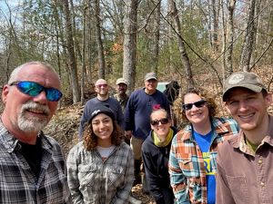 A selfie shot of eight members of PA/DE staff posing together at the edge of the woods at Tannersville Cranberry Bog Preserve.