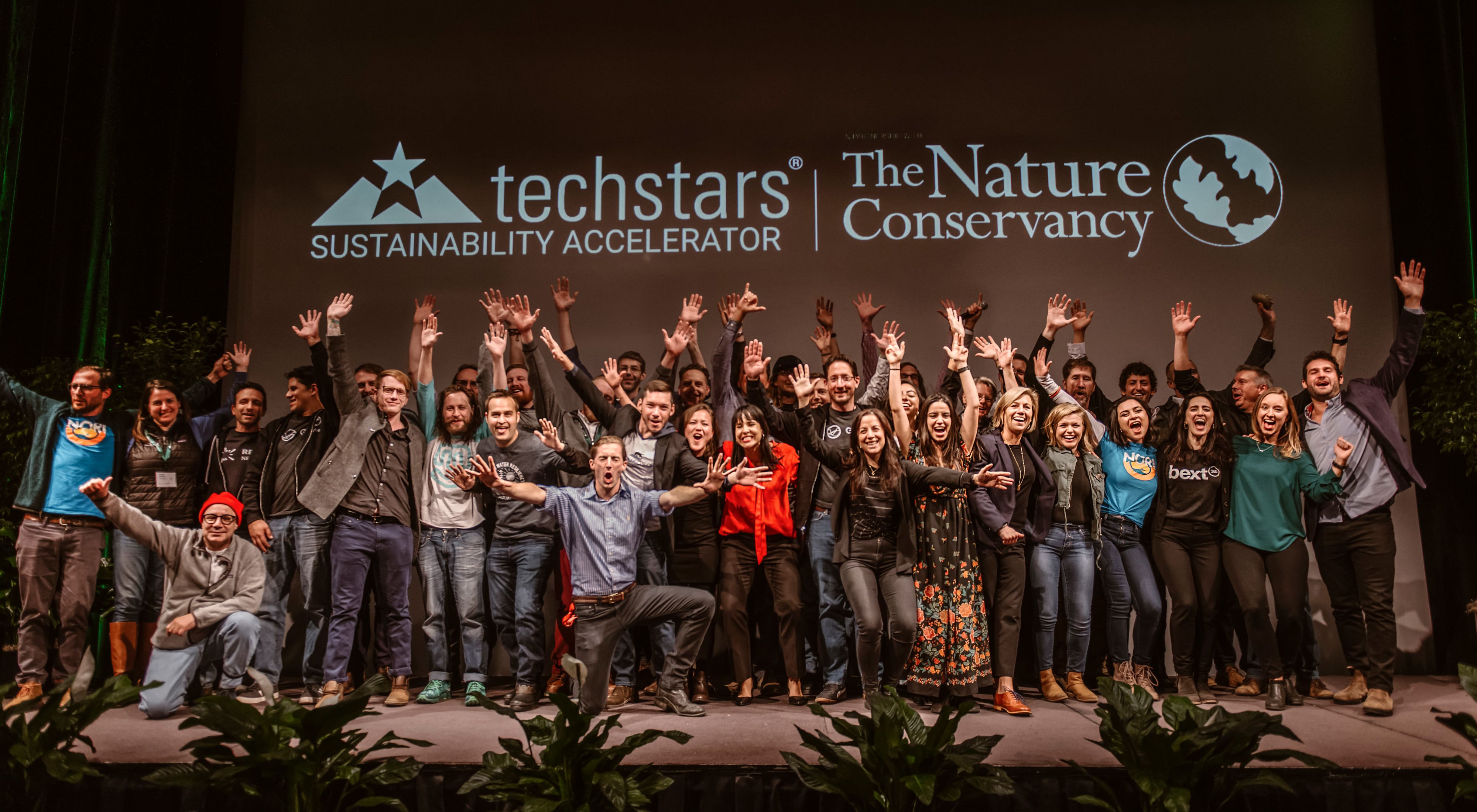 a large group of people on a stage holding their arms outstretched, with techstars and TNC logos behind them
