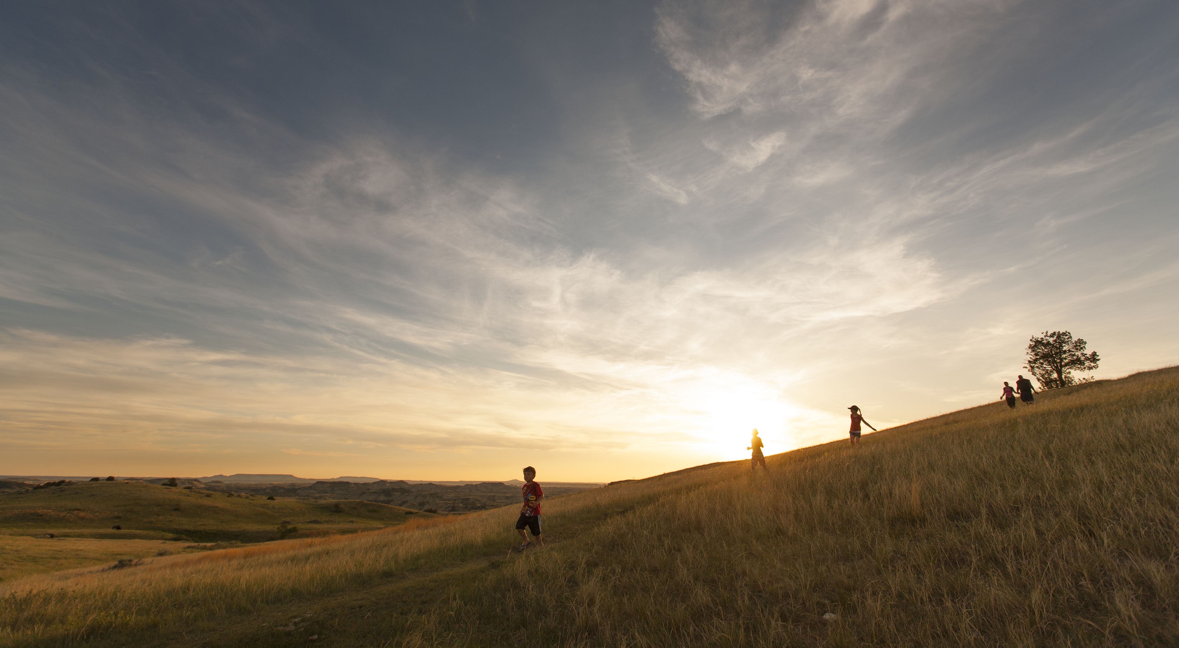 Kids run down a hillside trail ahead of their parents in with rolling grassy hills in the distance and the sun setting behind them.