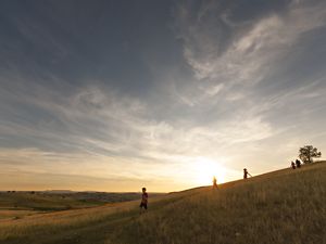 Landscape view looking across grassland hills as several people walk downhill with the sun setting in the background.