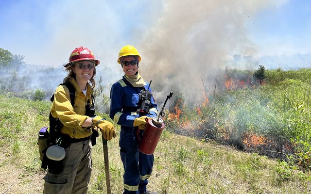 Two women wearing hard hats carry a drip torch while standing in front of a burning landscape.