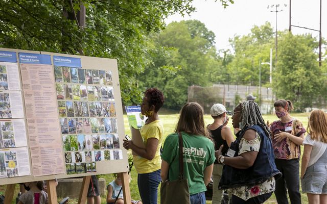 A group of people looking at a board with pictures of trees while standing in a park. 
