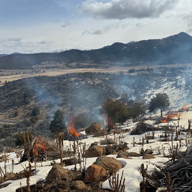 View looking across a mountainous landscape with several piles of tree debris burning in the foreground.