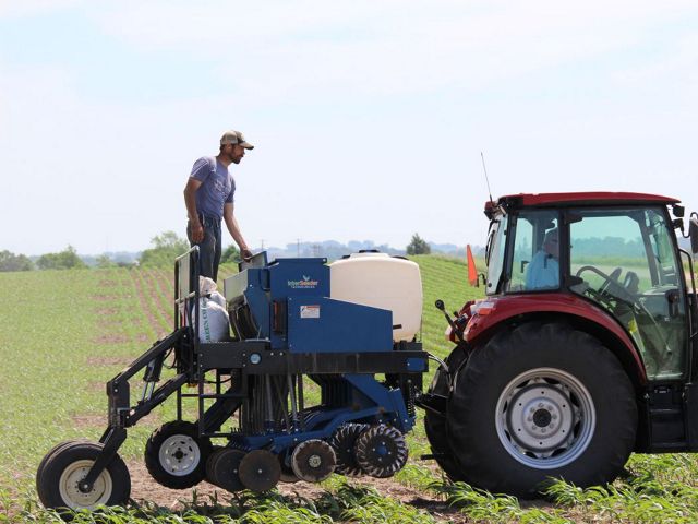 A man stands on top of an agricultural seeder that is attached to a tractor.
