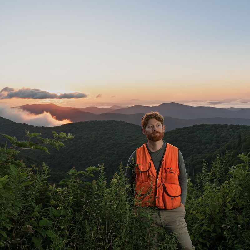 a man in an orange vest stands amid shrubs overlooking mountains in the background during sunset.