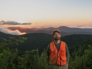 a man in an orange vest stands amid shrubs overlooking mountains in the background during sunset.