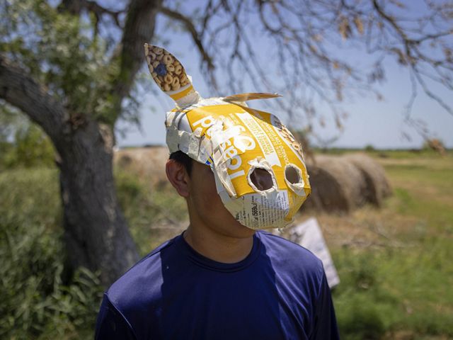 child in blue shirt has an animal-like paper mache mask with ears and snout over his face, haybales and tree in background