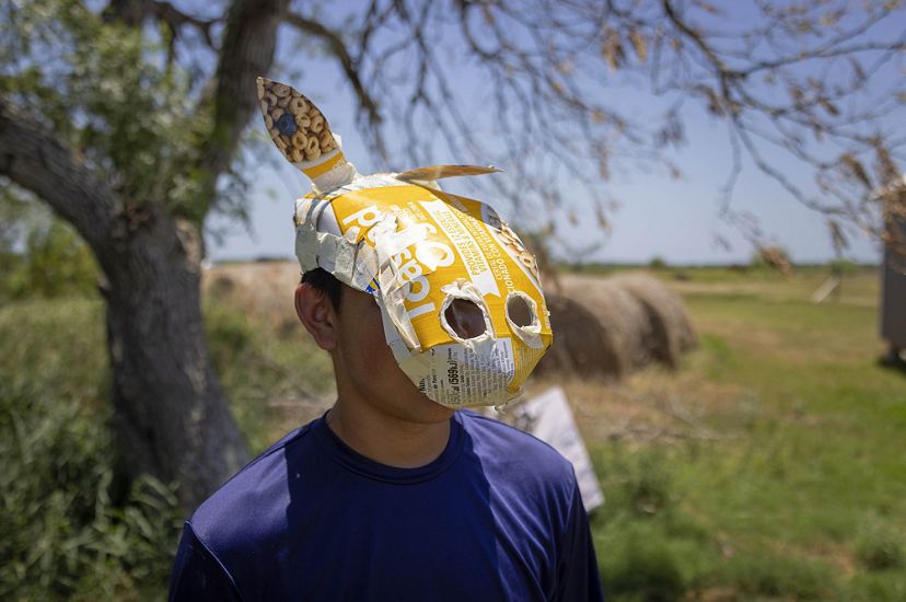 child in blue shirt has an animal-like paper mache mask with ears and snout over his face, haybales and tree in background