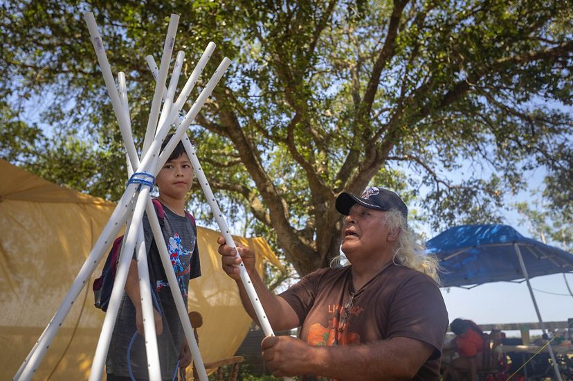 an older man and a young boy seen mid-movement, arranging white poles upright in a tepee formation