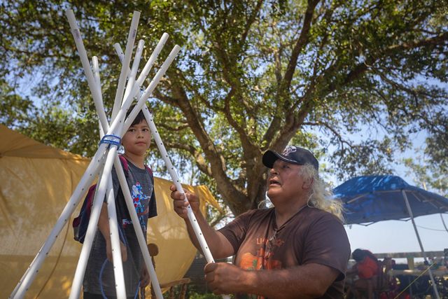 an older man and a young boy seen mid-movement, arranging white poles upright in a tepee formation