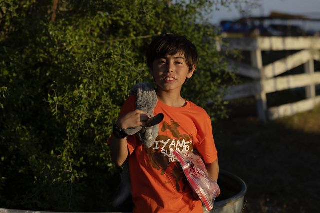 young boy faces camera, half in shadow, the pelt of a rabbit hangs over his shoulder and he holds a plastic bag with meat in it