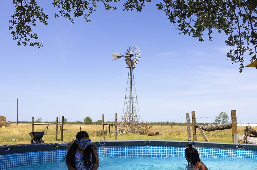 3 kids play in an above ground poll in the shade of a tree whose limbs arch at the border of the frame. Windmill and ranch fencing in background