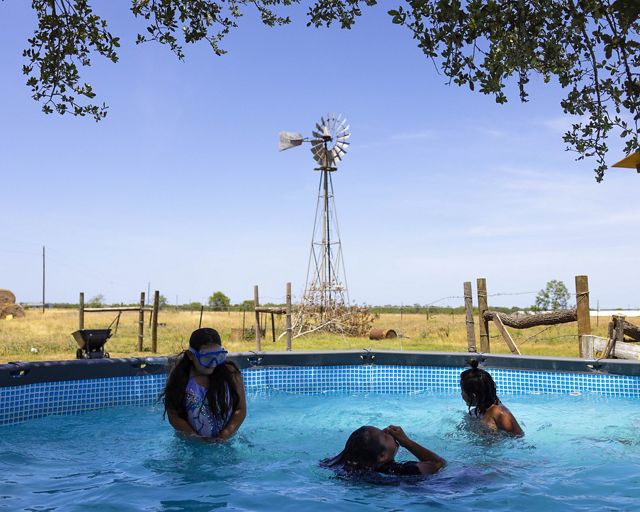 3 kids play in an above ground poll in the shade of a tree whose limbs arch at the border of the frame. Windmill and ranch fencing in background