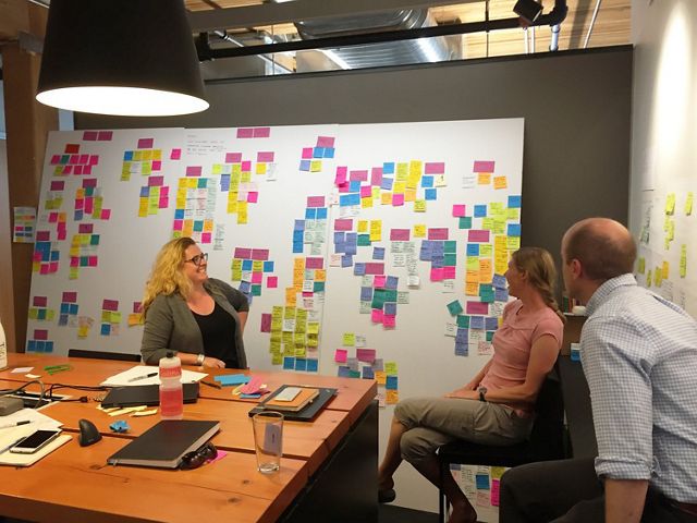 Three people sit in a small room with a whiteboard covered in post-it notes.