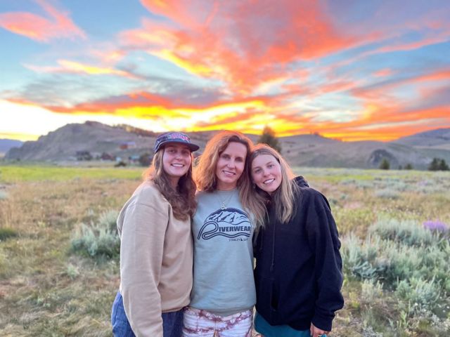 Three women stand in front of a mountain range at sunset.