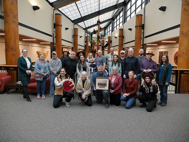 A group of people pose together following a ceremony. The person in the center of the group holds a framed photo. Another person kneels and holds a drum.