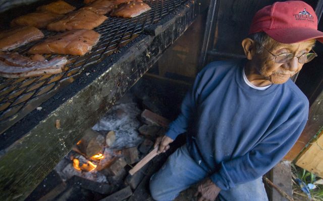 A man sits near a small fire and cooks salmon.