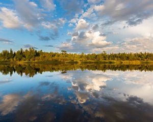 A pale blue sky with clouds is reflected in a still lake that is bordered by trees.