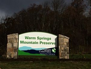 A large sign held up by two stone pillars welcomes visitors. The sign reads Warm Springs Mountain Preserve. At the bottom is stylized mountain graphic with the TNC logo in the corner.
