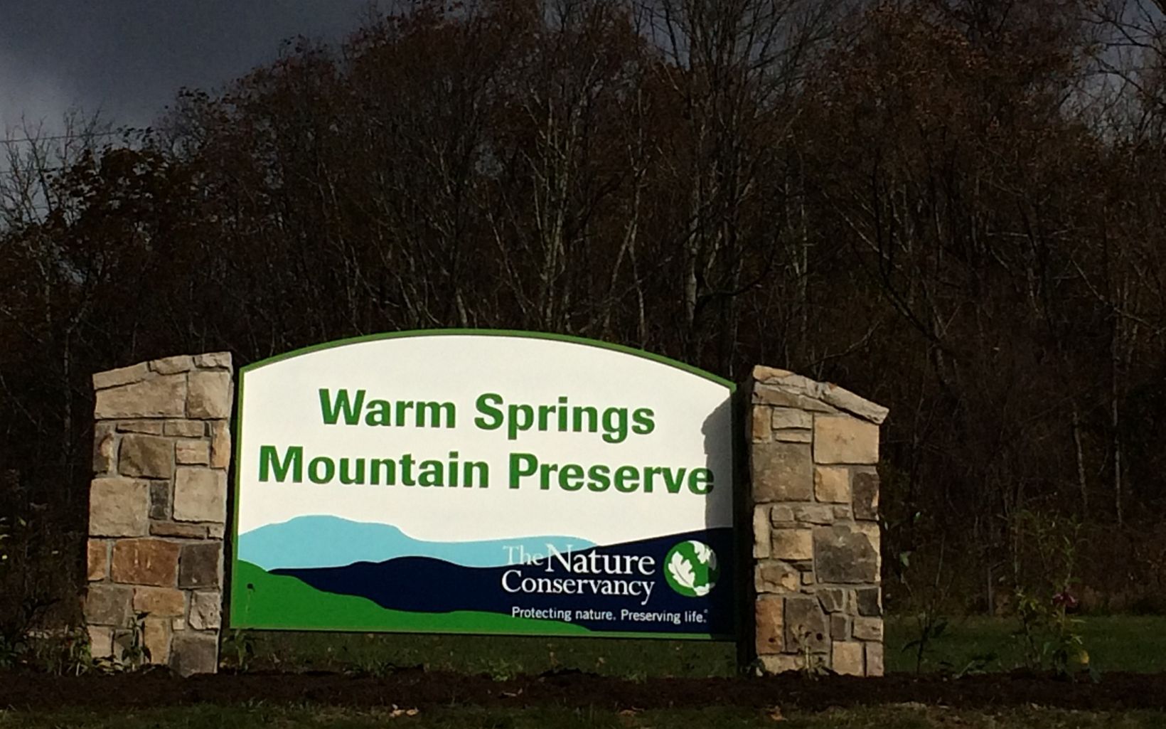 A large sign held up by two stone pillars welcomes visitors. The sign reads Warm Springs Mountain Preserve. At the bottom is stylized mountain graphic with the TNC logo in the corner.