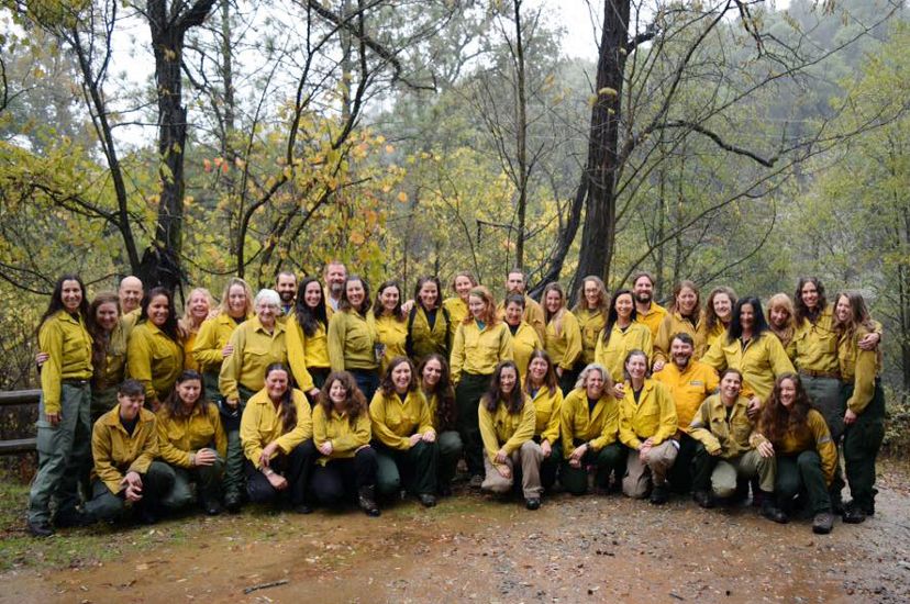 Group photo of several dozen women taking during a fire training exchange. The women are wearing yellow fire gear and posing in a clearing in a forest.