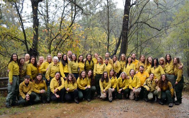 Group photo of several dozen women taking during a fire training exchange. The women are wearing yellow fire gear and posing in a clearing in a forest.