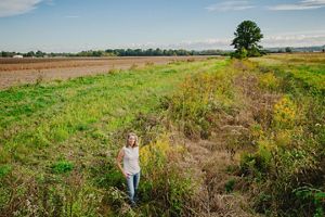 Woman stands in ditch alongside ag field.