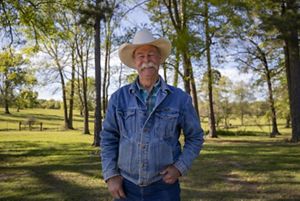 A man in a white cowboy hat stands surrounded by green trees.