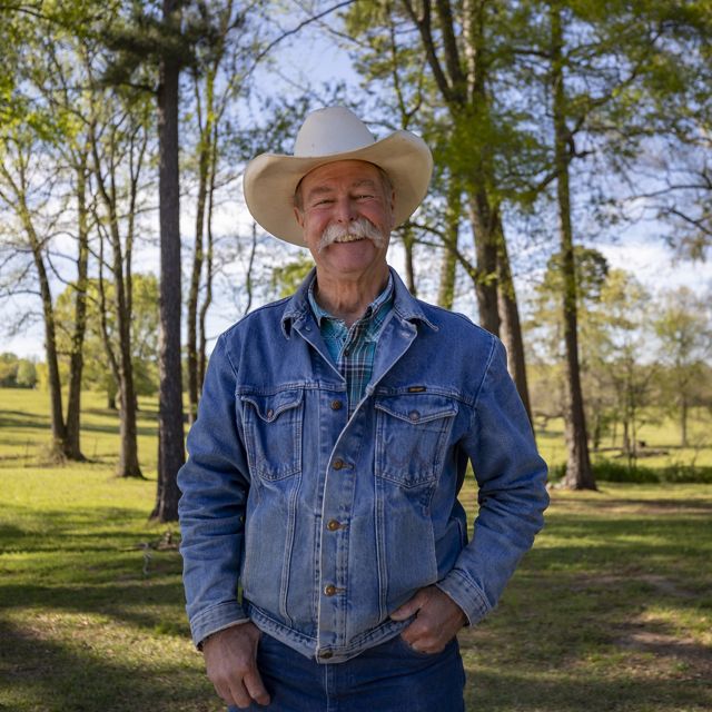A man in a white cowboy hat stands surrounded by green trees.