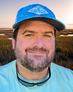 A headshot of Aaron Bunch, showing a man with cap in front of coastal wetlands.