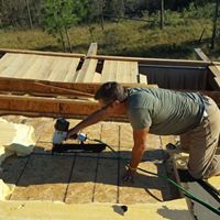 A man kneels on a roof and uses a power tool to attach boards.