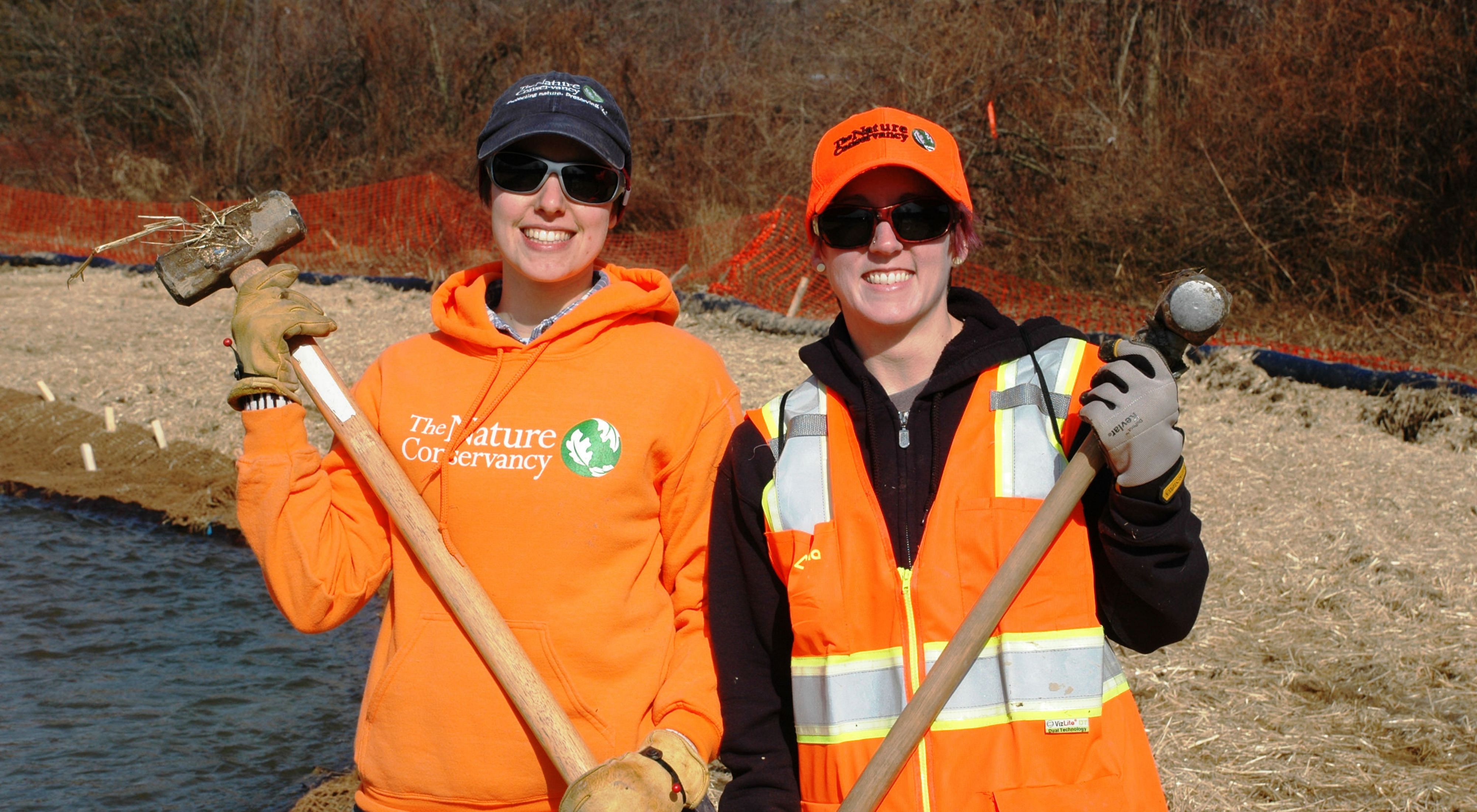 Two smiling women pose together during a volunteer event. They are both wearing orange hats and vests and holding sledge hammers. Sedge and straw cover the ground behind them.