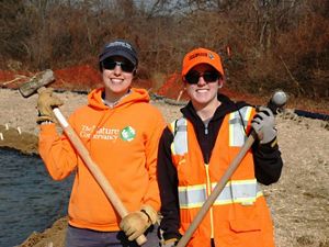 Two smiling women pose together during a volunteer event. They are both wearing orange hats and vests and holding sledge hammers. Sedge and straw cover the ground behind them.