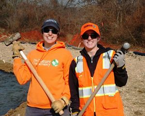 Two smiling women pose together during a volunteer event. They are both wearing orange hats and vests and holding sledge hammers. Sedge and straw cover the ground behind them.