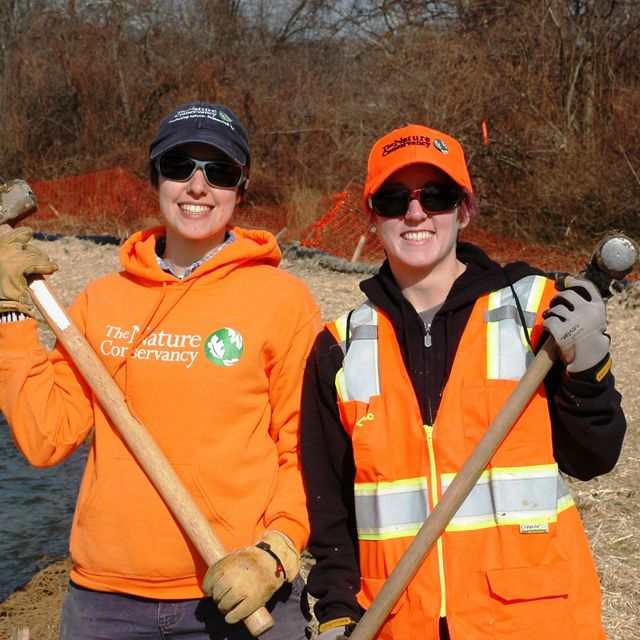 Two smiling women wearing orange vests and caps and holding sledgehammers.