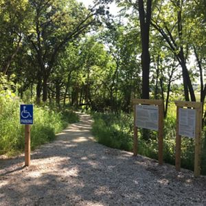 A dirt path winds into a wooded area; trail signage is in the right foreground.
