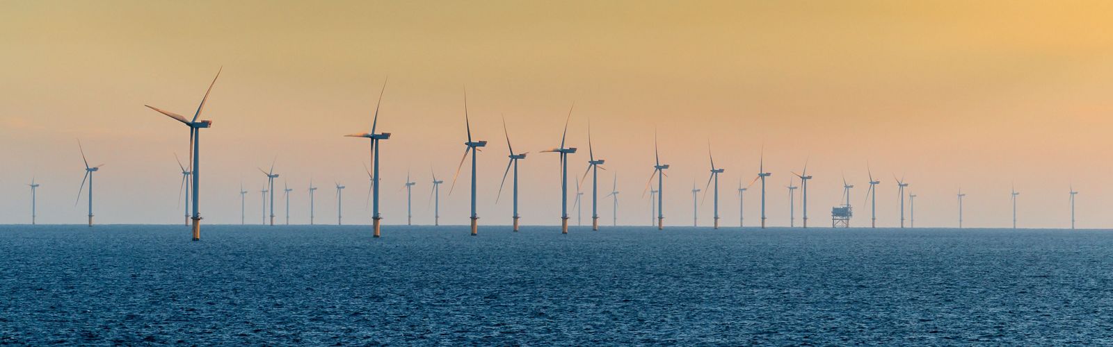 The sun sets over an ocean with several wind turbines.