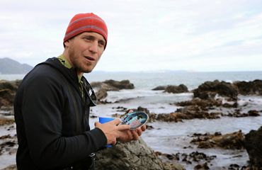 Adam Starke holds a seashell while standing on a rocky coast as he works to collect eelgrass.