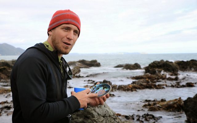 Adam Starke holds a seashell while standing on a rocky coast as he works to collect eelgrass.