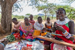 A group of women sit beneath a tree, singing as they assemble beaded jewelry.