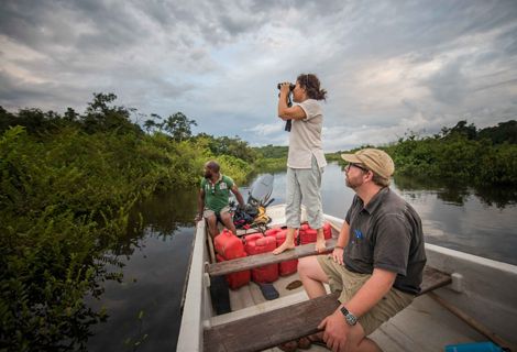 Three people on a boat on a body of water, one stands with binoculars looking out into a lush vegetated area.