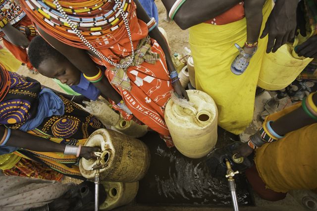 A group of women and children in colorful clothing gather around a well to collect water in large jugs.