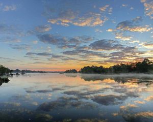 The sun rises over a placid river, creating reflections of orange sky and puffy clouds on the still water's surface.