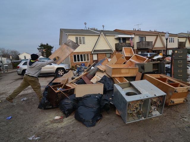 Man tosses broken cabinets onto a pile of destroyed appliances and furniture on Staten Island residential street with two-story, paneled homes and cars in the background.