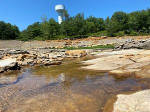 A stream runs through a rocky channel, with trees and a water tower in the background.