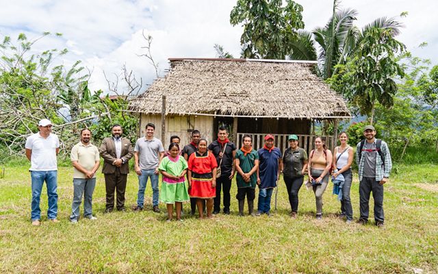 A group of people stand in a line in front of a small, grass-roofed building and trees.