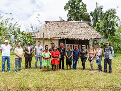 A group of people stand in a line in front of a small, grass-roofed building and trees.