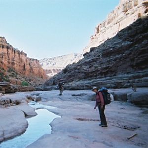 Two people with hiking backpacks stand in a valley on a sunny day with a small stream of water going through it