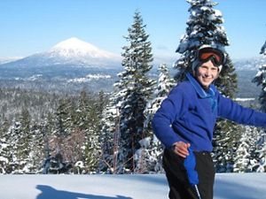 A woman wearing a blue winter coat and sky goggles stands and smiles with pine trees covered in a snow and a snow-capped mountain behind her.