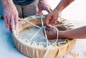Closeup of hands tying an animal hide around a round base, forming a drum.